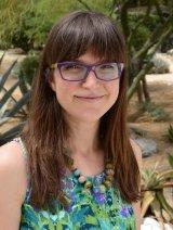 Woman with brown hair, wearing glasses, and wearing a green shirt and beaded necklace standing outdoors.